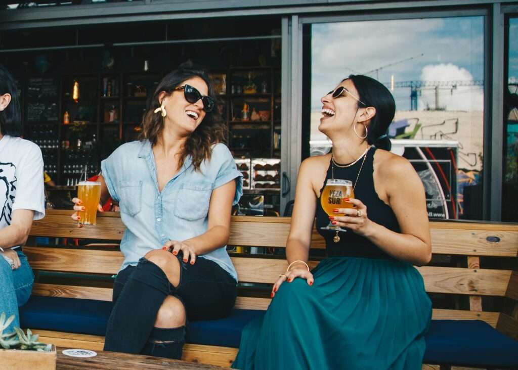 Two women laughing and enjoying drinks outdoors at a trendy bar, creating a vibrant and social atmosphere.