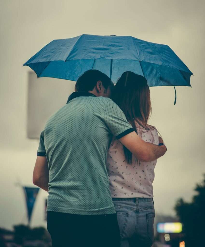 A couple embraces under a blue umbrella in a rainy street scene, evoking love and warmth.