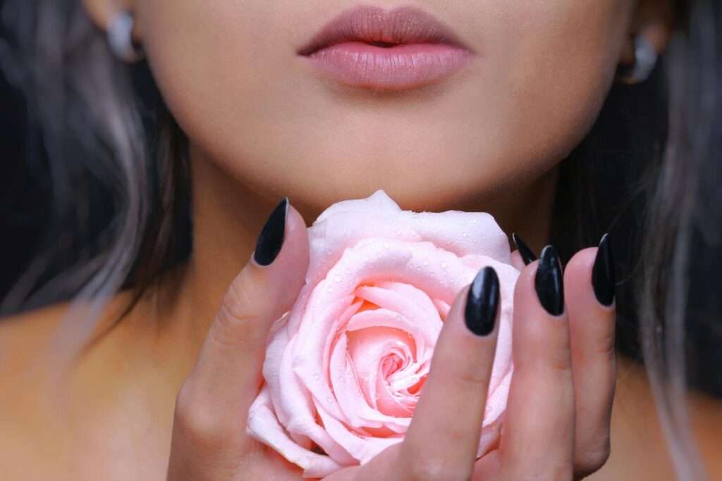 A close-up of a woman with a pink rose, highlighting her lips and black nails.