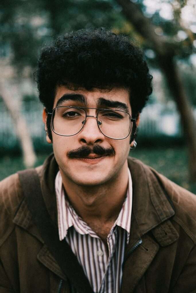 Young man with curly hair and glasses making a funny face in an outdoor setting.