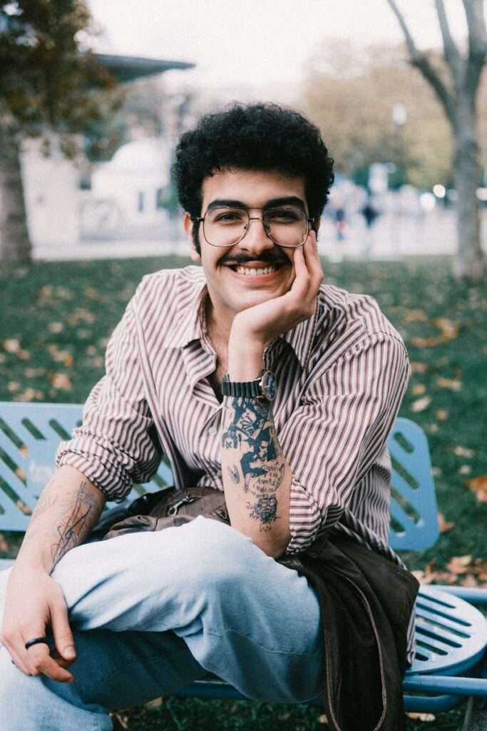 Casual portrait of a smiling adult male with tattoos sitting on a park bench during fall.