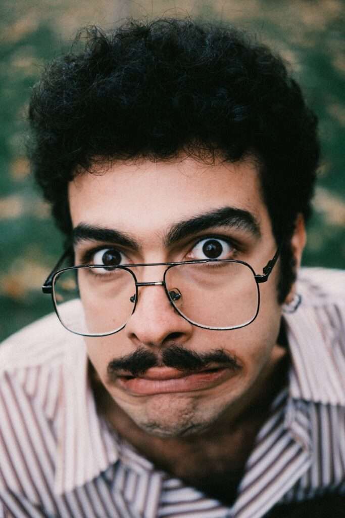 Close-up of a playful man with big glasses and a mustache outdoors.