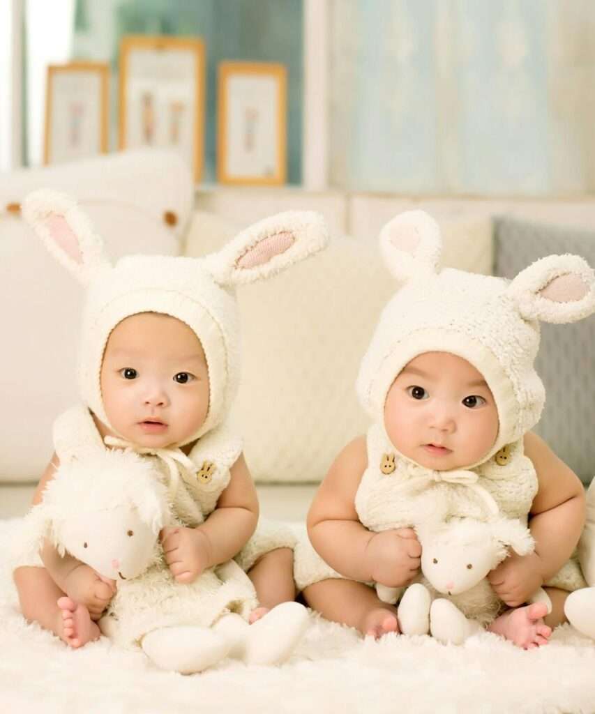 Twin babies wearing cute bunny outfits with plush toys indoors.