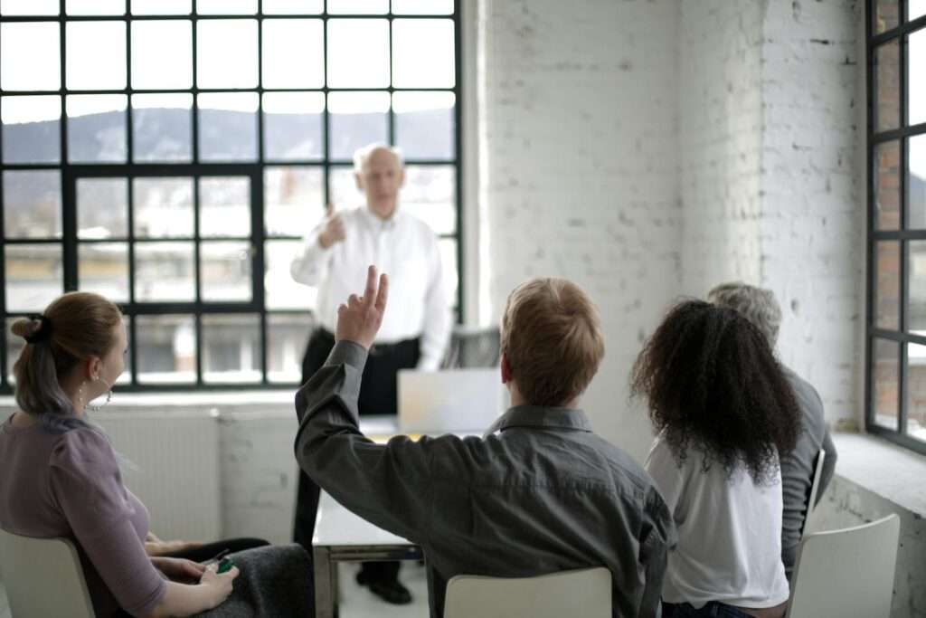 Group of adults in a discussion, with one person raising a hand in a bright office space.