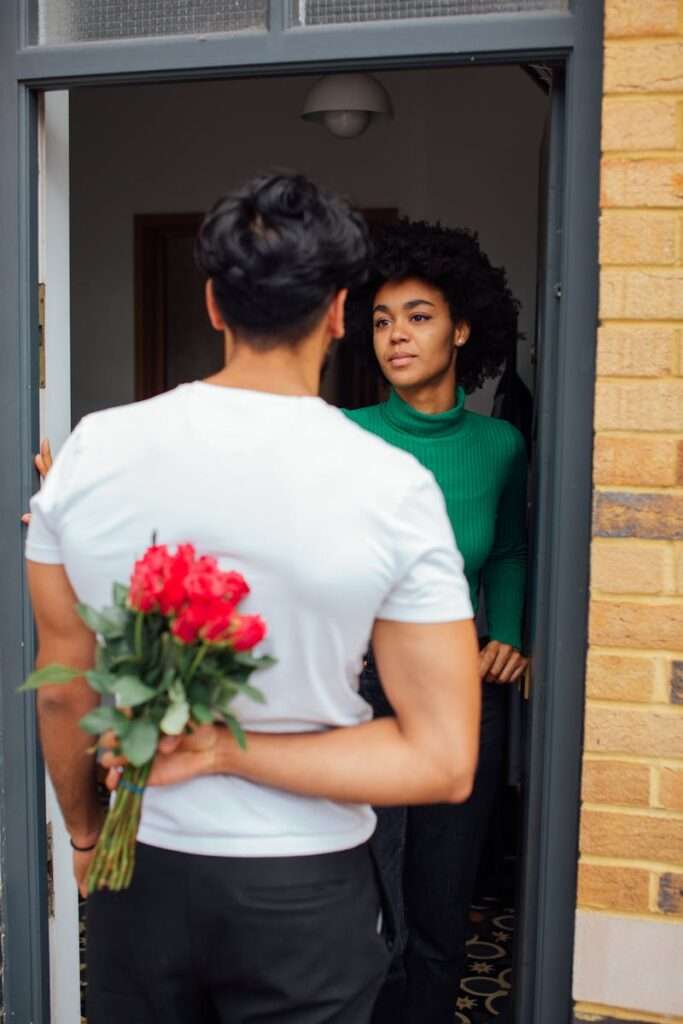 A man surprises a woman with red roses at her doorstep, symbolizing romance.