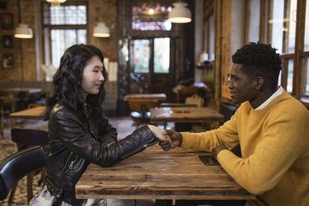 A diverse man and woman sit in a café, shaking hands and smiling warmly.