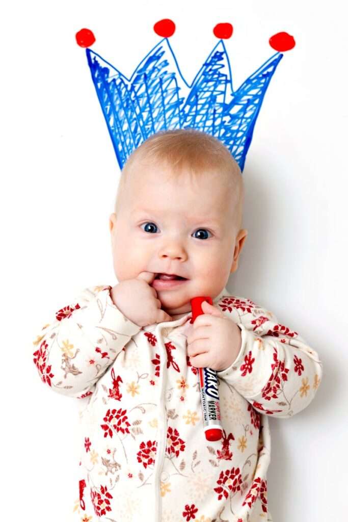 Cute baby wearing patterned clothing, holding a marker with a crown drawn above, exhibiting joy and innocence.