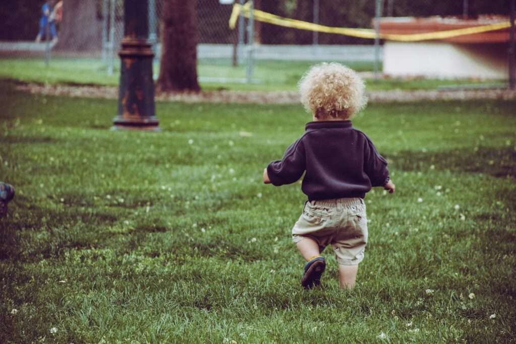 A curly-haired toddler joyfully walks in a grassy park, capturing childhood innocence.