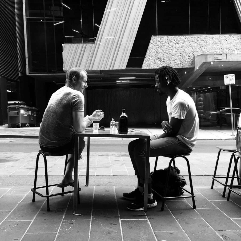 Black and white photo of two men chatting outside on stools in South Yarra, Melbourne.