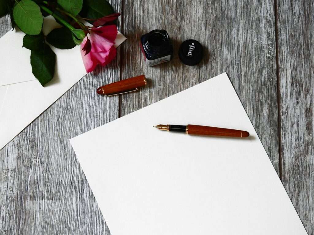 Flat lay image of a rose, fountain pen, and paper on a rustic wooden table.