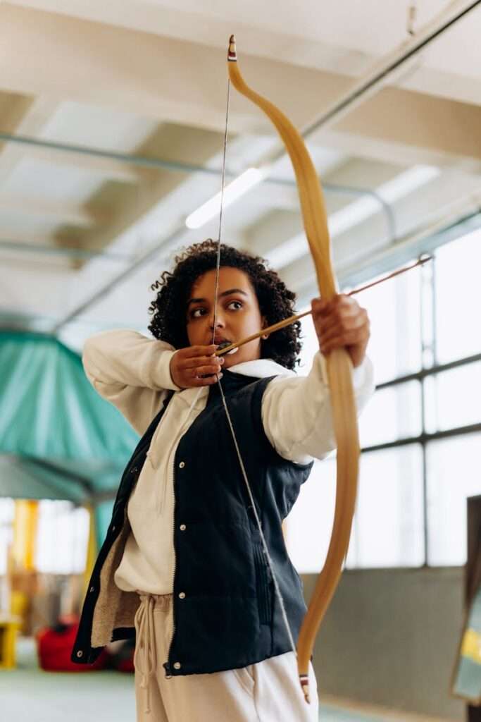 A young woman concentrating as she aims her bow and arrow, practicing archery indoors.